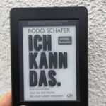 Das Buchcover von "Ich kann das." von Bodo Schäfer auf einem Kindle, gehalten von einer Hand vor einer weißen Raufasertapete.