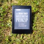 Das Buchcover von Das Brian Tracy Maximum Prinzip auf einem Kindle, der im Sommer auf einer Wiese im Gras liegt.