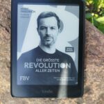 Das Buchcover von 'Die grösste Revolution aller Zeiten' von Marc Friedrich, angezeigt auf einem Kindle, der auf einem Stein liegt.