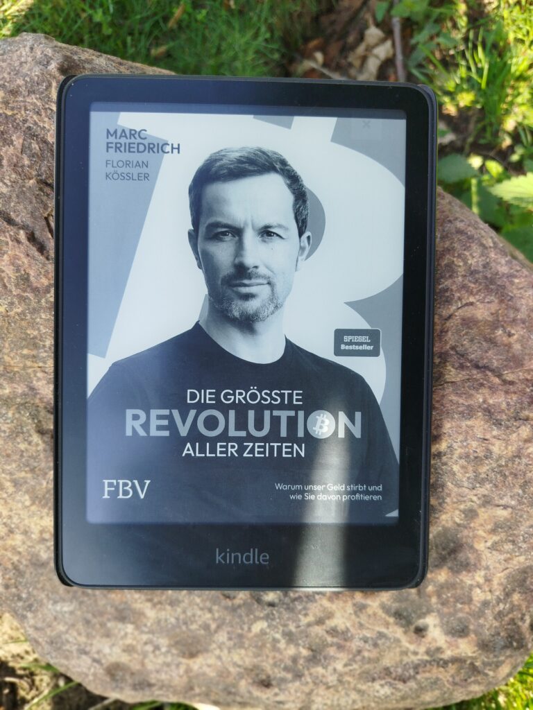 Das Buchcover von 'Die grösste Revolution aller Zeiten' von Marc Friedrich, angezeigt auf einem Kindle, der auf einem Stein liegt.