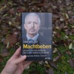 Das Buch 'Machtbeben' von Dirk Müller in einer Hand gehalten, draußen vor einem Hintergrund aus herbstlichem Laub.