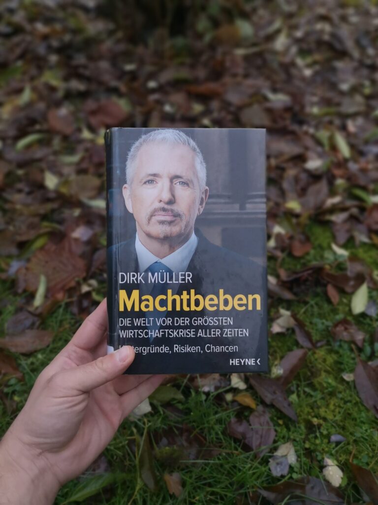 Das Buch 'Machtbeben' von Dirk Müller in einer Hand gehalten, draußen vor einem Hintergrund aus herbstlichem Laub.