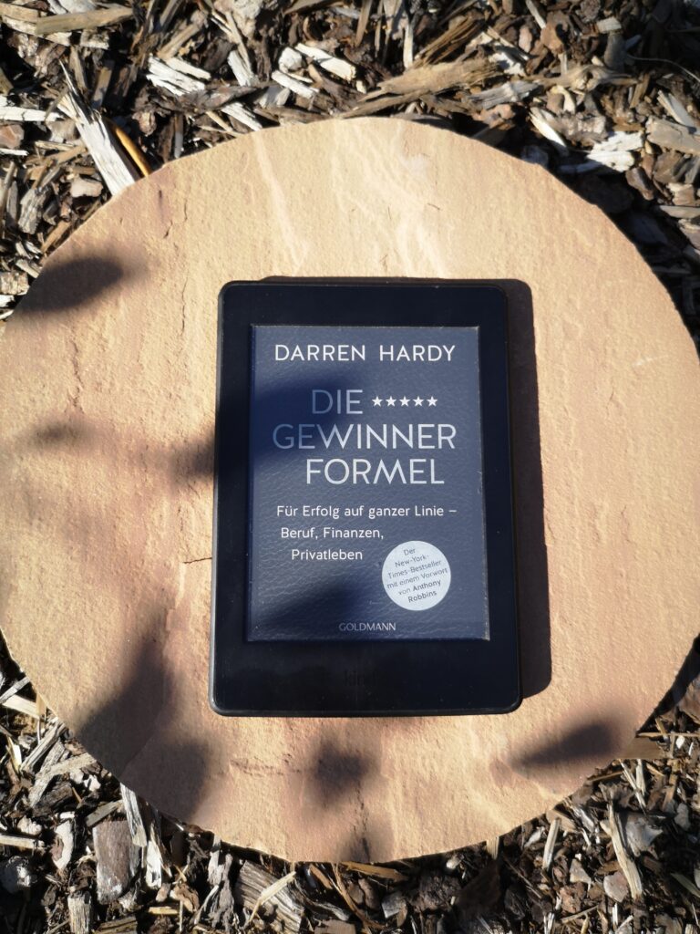 Das Buchcover von Die Gewinner-Formel von Darren Hardy auf einem Kindle, der draußen auf einem Stein im Garten liegt.