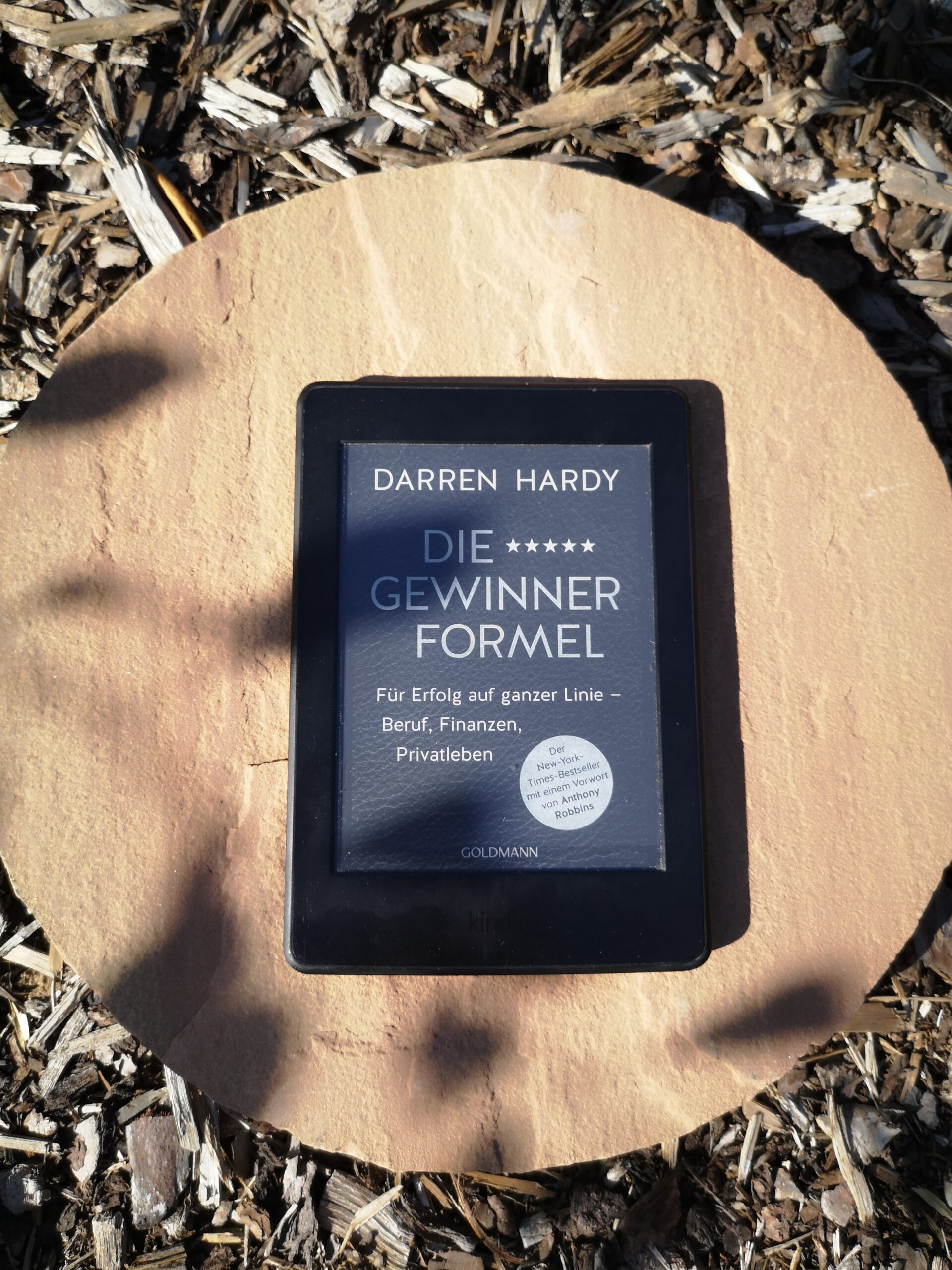 Das Buchcover von Die Gewinner-Formel von Darren Hardy auf einem Kindle, der draußen auf einem Stein im Garten liegt.