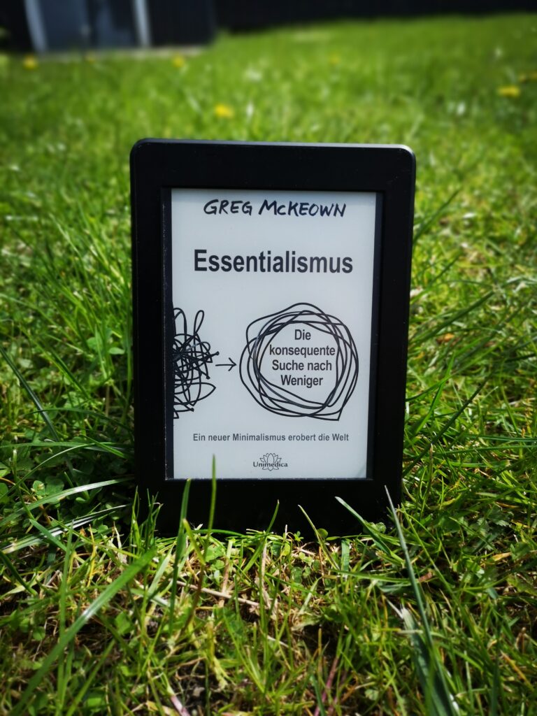 Ein Kindle E-Reader zeigt das Cover des Buches Essentialismus von Greg McKeown, das in einer grünen Wiese steht.