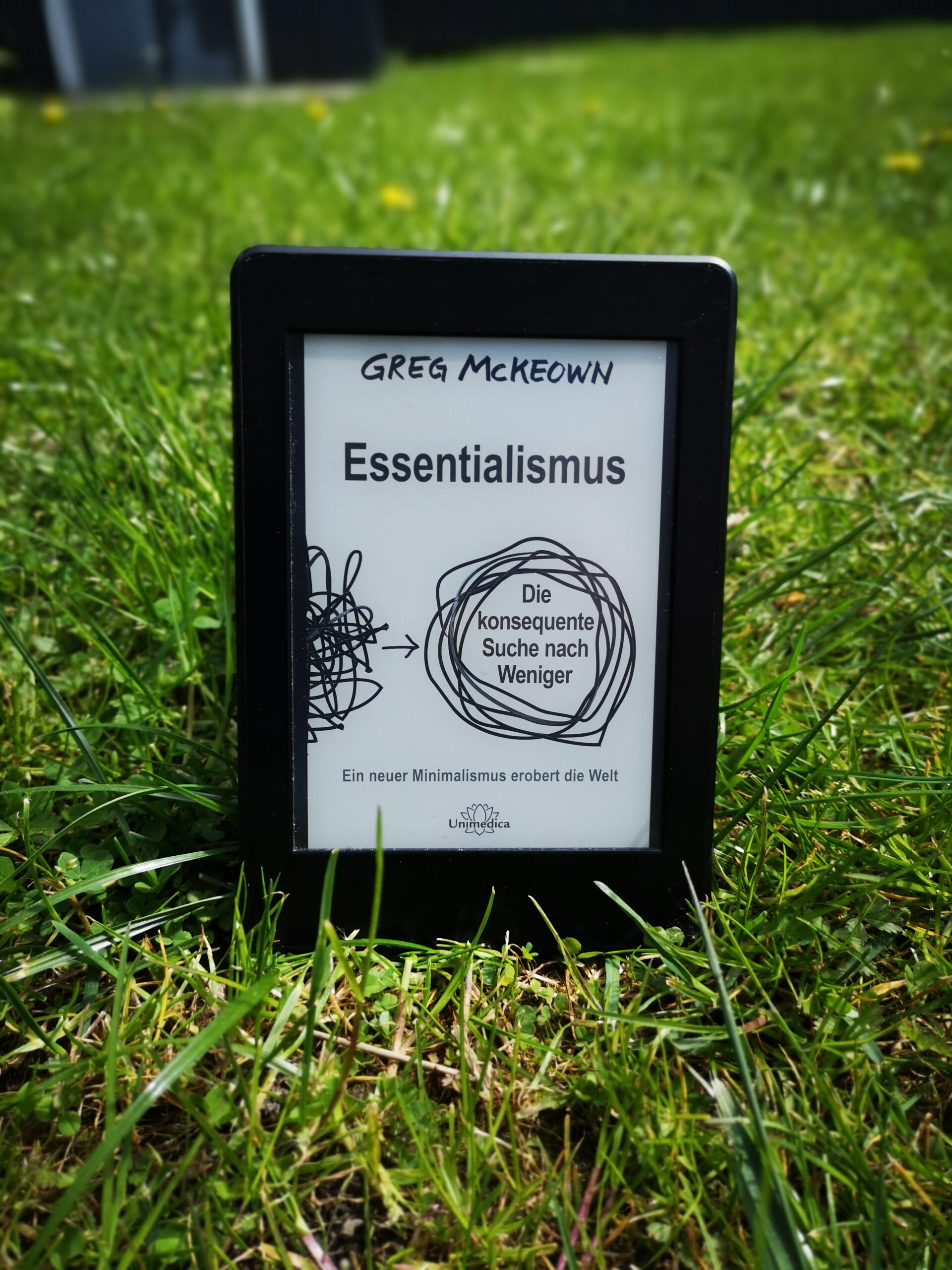 Ein Kindle E-Reader zeigt das Cover des Buches Essentialismus von Greg McKeown, das in einer grünen Wiese steht.