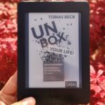 Ein Kindle E-Reader zeigt das Cover von 'Unbox Your Life!' von Tobias Beck, gehalten in einer Hand vor einem Hintergrund aus runden, roten Blättern (Ahorn).