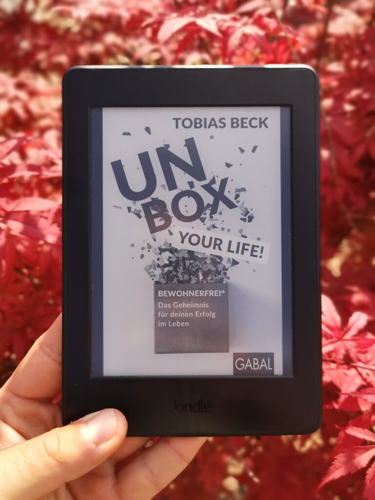 Ein Kindle E-Reader zeigt das Cover von 'Unbox Your Life!' von Tobias Beck, gehalten in einer Hand vor einem Hintergrund aus runden, roten Blättern (Ahorn).