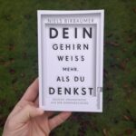 Das Buch "Dein Gehirn weiß mehr als du denkst" mit Rasen im Hintergrund durch eine Hand gehalten